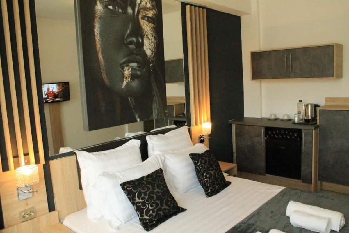 A modern hotel room with a large bed, a flat-screen TV, a small kitchen area, and a large artwork on the wall.