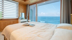 A bedroom with a large bed, a bedside table, a lamp, and a view of the ocean.