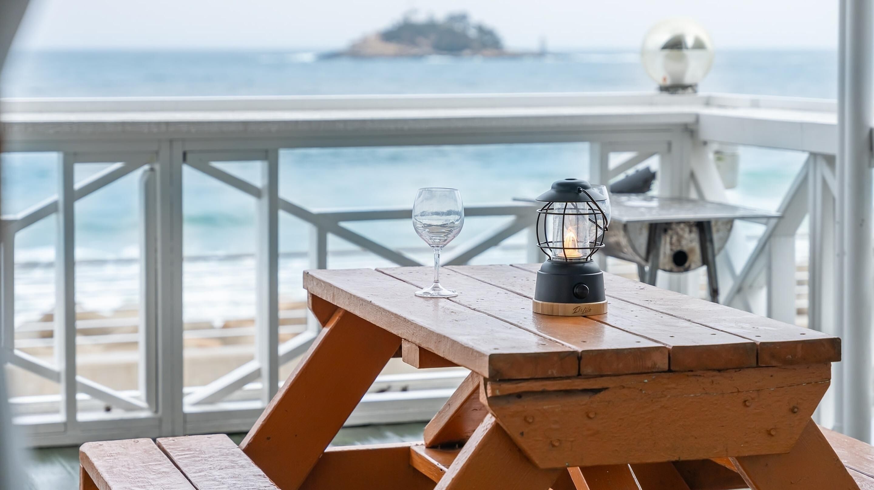 A wooden table with a glass of water and a lantern on a deck with a railing, overlooking the sea.