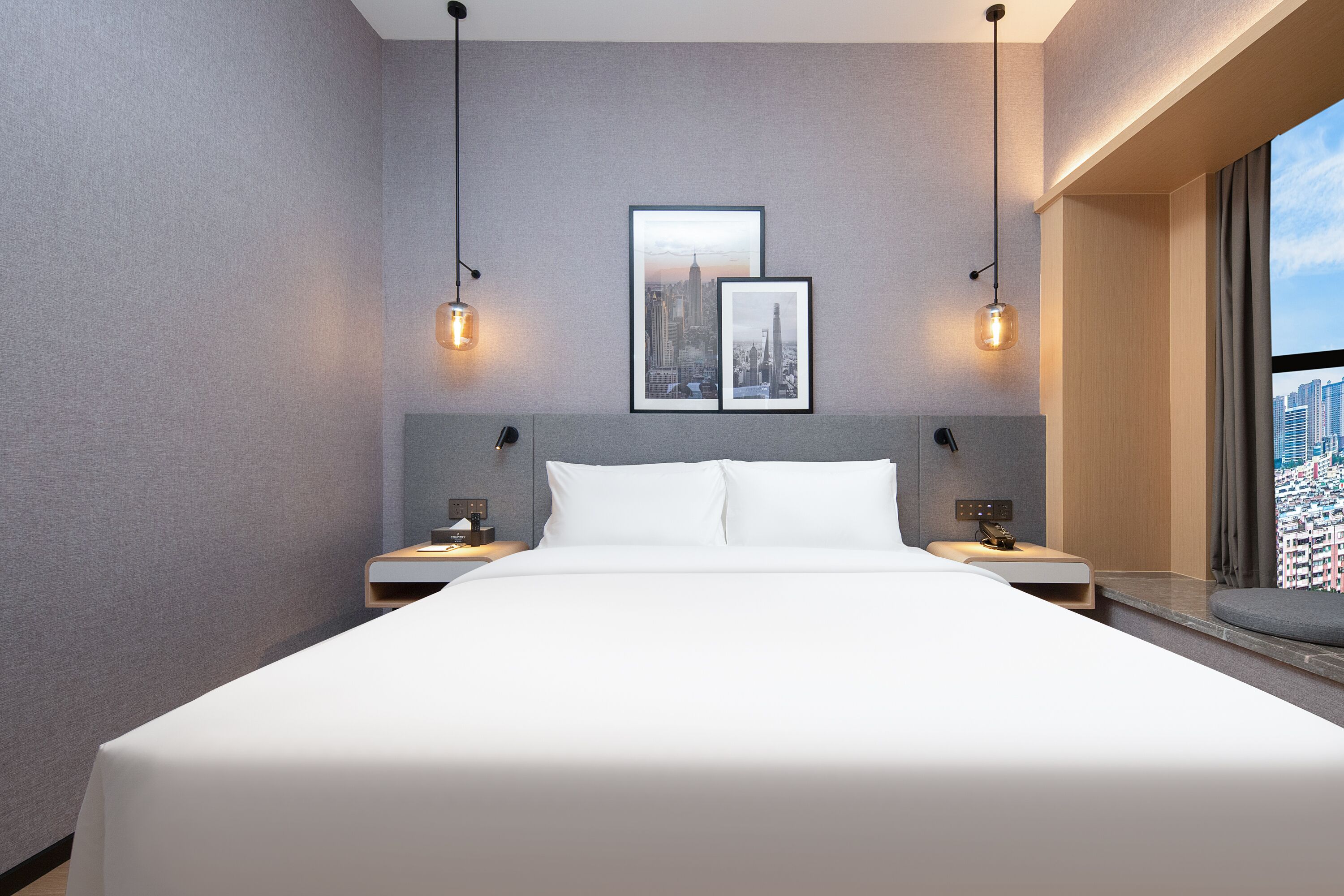 Una habitación de hotel moderna con una cama grande, dos mesitas de noche, vistas a la ciudad e iluminación colgante.