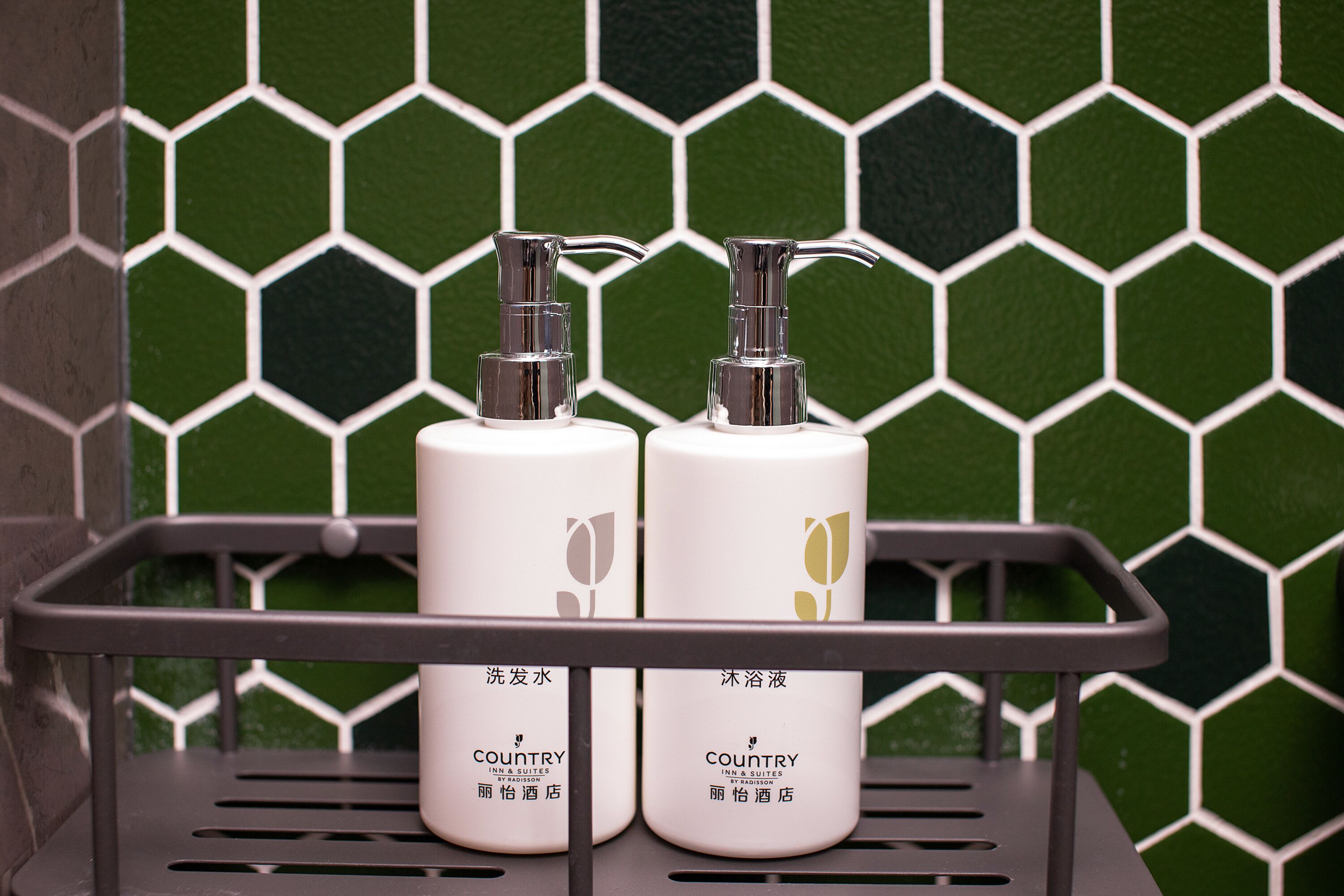 Dos botellas de artículos de tocador del hotel Country sobre un estante de metal frente a una pared de azulejos hexagonales verdes.