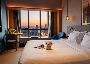 A hotel room with a large bed, a desk with a chair, a window with a city view, and a hanging light fixture.
