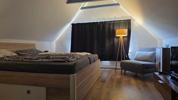 A bedroom with a bed, a chair, a floor lamp, and a TV stand. - Dachgeschosswohnung - Erholung mit Waldblick (Mustin)