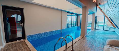 A modern indoor swimming pool with a wooden floor, a glass barrier, and a skylight.