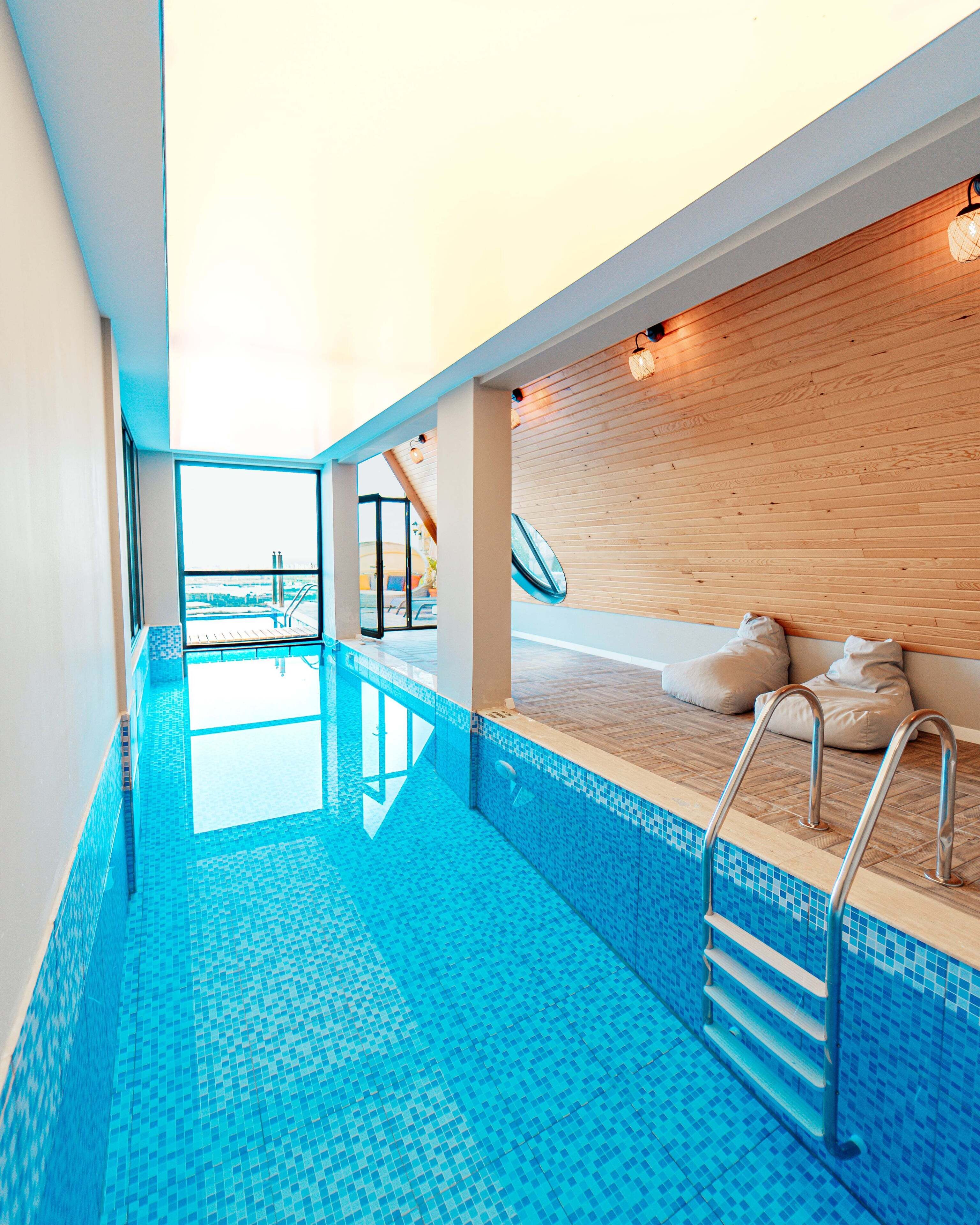 A modern indoor swimming pool with clear blue tiles, a wooden deck area, and a view of the cityscape.
