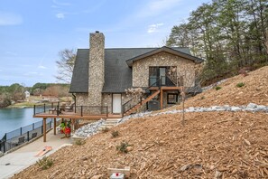 Una casa de piedra con balcón que da a un lago, una terraza y una escalera que lleva a la entrada principal.