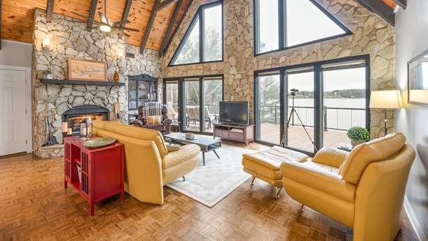 A spacious living room with a stone fireplace, wooden ceiling beams, and large windows offering a view of the outdoors.