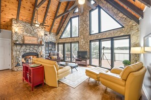 Un salón amplio con chimenea de piedra, vigas de madera en el techo y grandes ventanales con vistas al exterior.