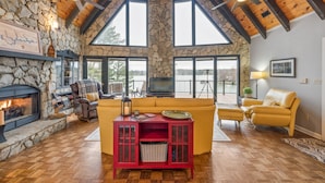 A cozy living room with a stone fireplace, wooden beams, large windows, and a yellow sofa.