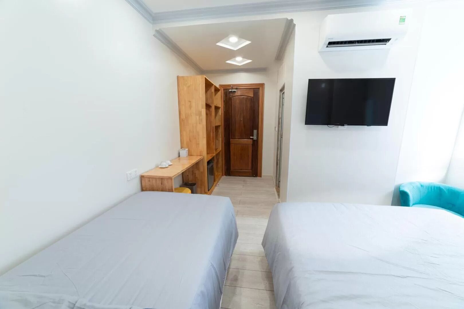 Una habitación pequeña y estrecha con dos camas individuales, un banco de madera, un televisor fijado a la pared y un equipo de aire acondicionado.