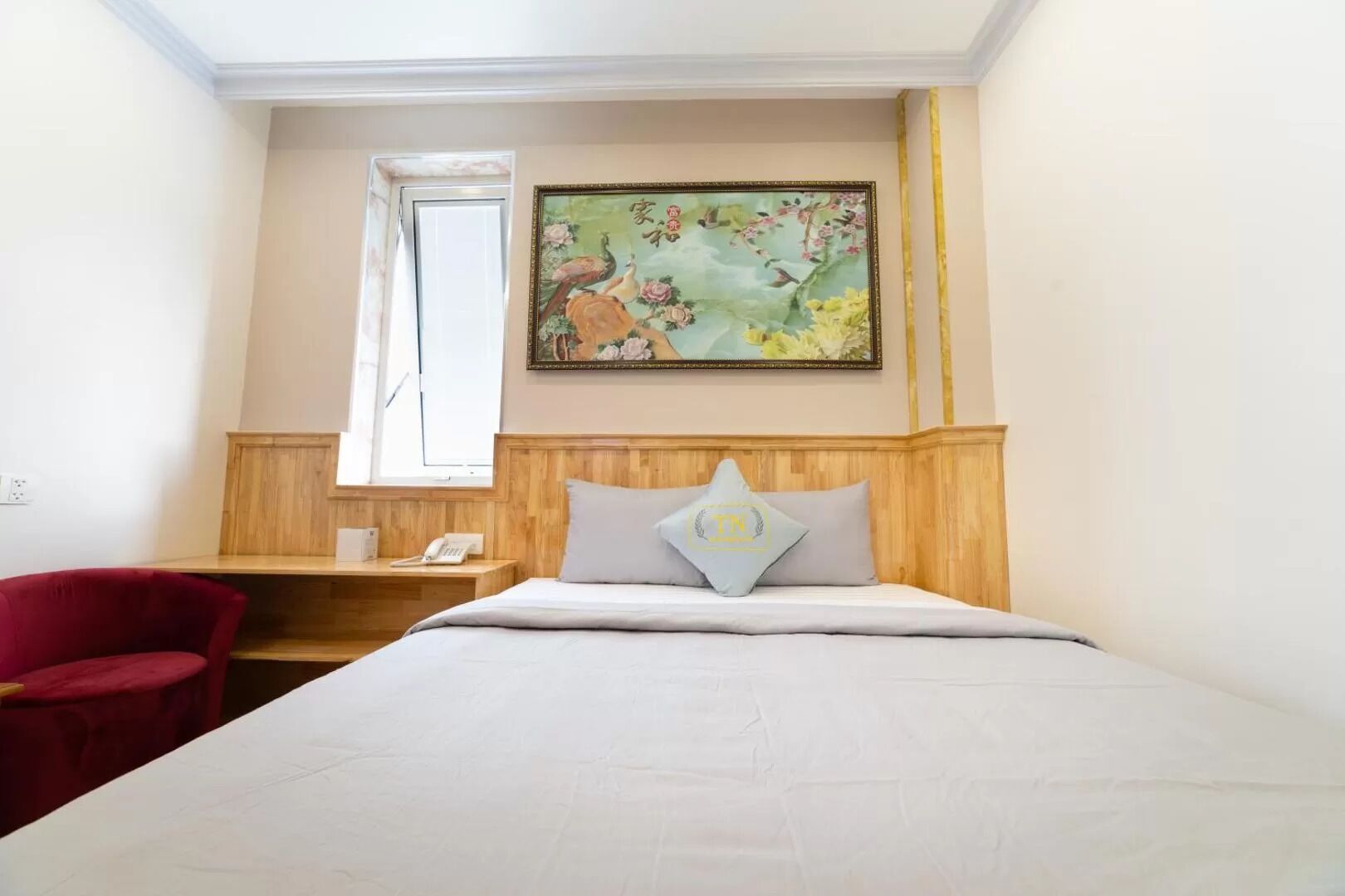 A hotel room with a bed, a red chair, a wooden nightstand with a phone, and a painting of birds and flowers on the wall.
