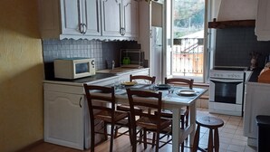 A compact kitchen with white cabinets, a microwave, a sink, and a stove. There is a dining table with chairs and a small stool. - Gite de Mon Clocher Appartement 3ième Étage Avec Balcon Proche du Mercantour (Roquebilliere)