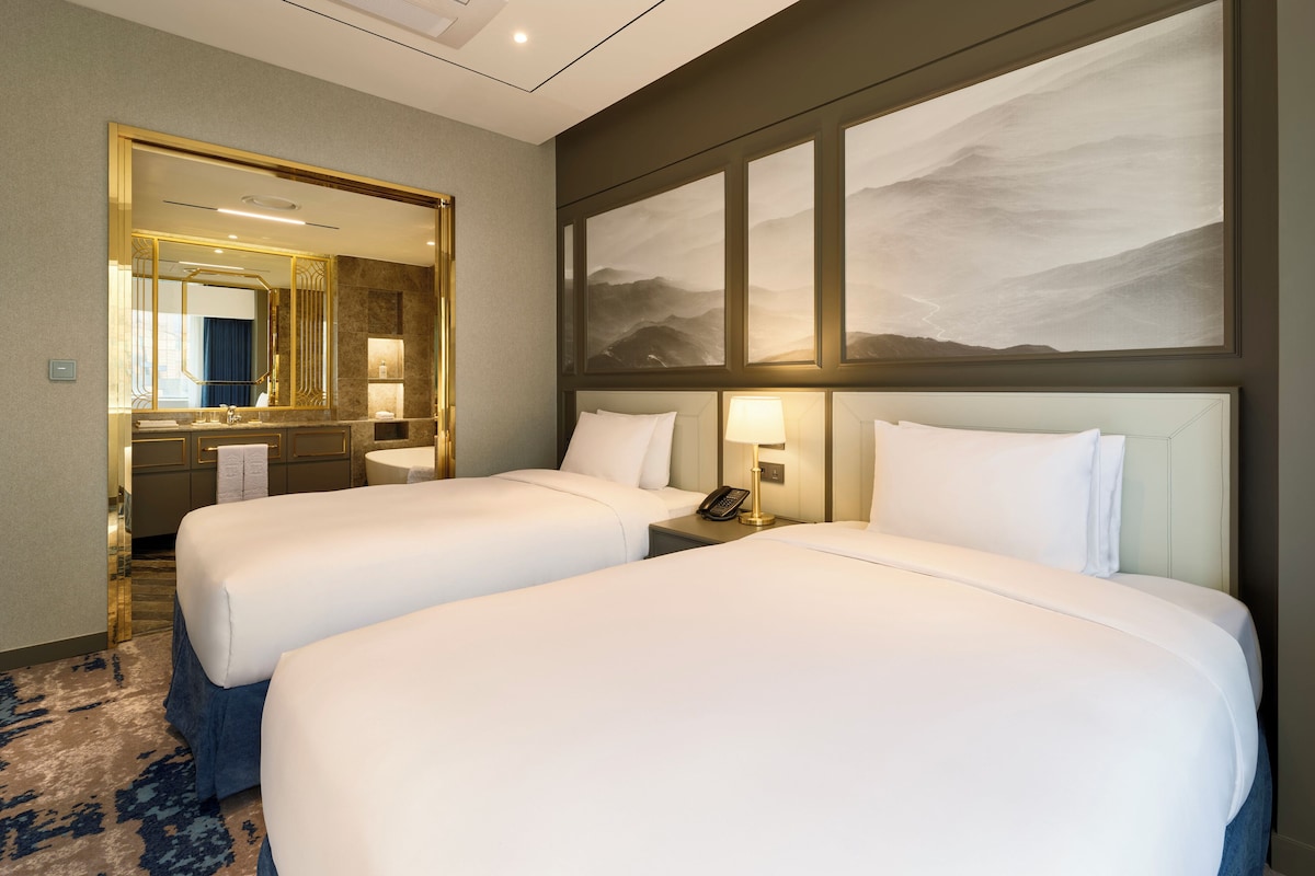 A hotel room with two beds, a large mirror, a painting, and a bathroom with a sink and towel rack.