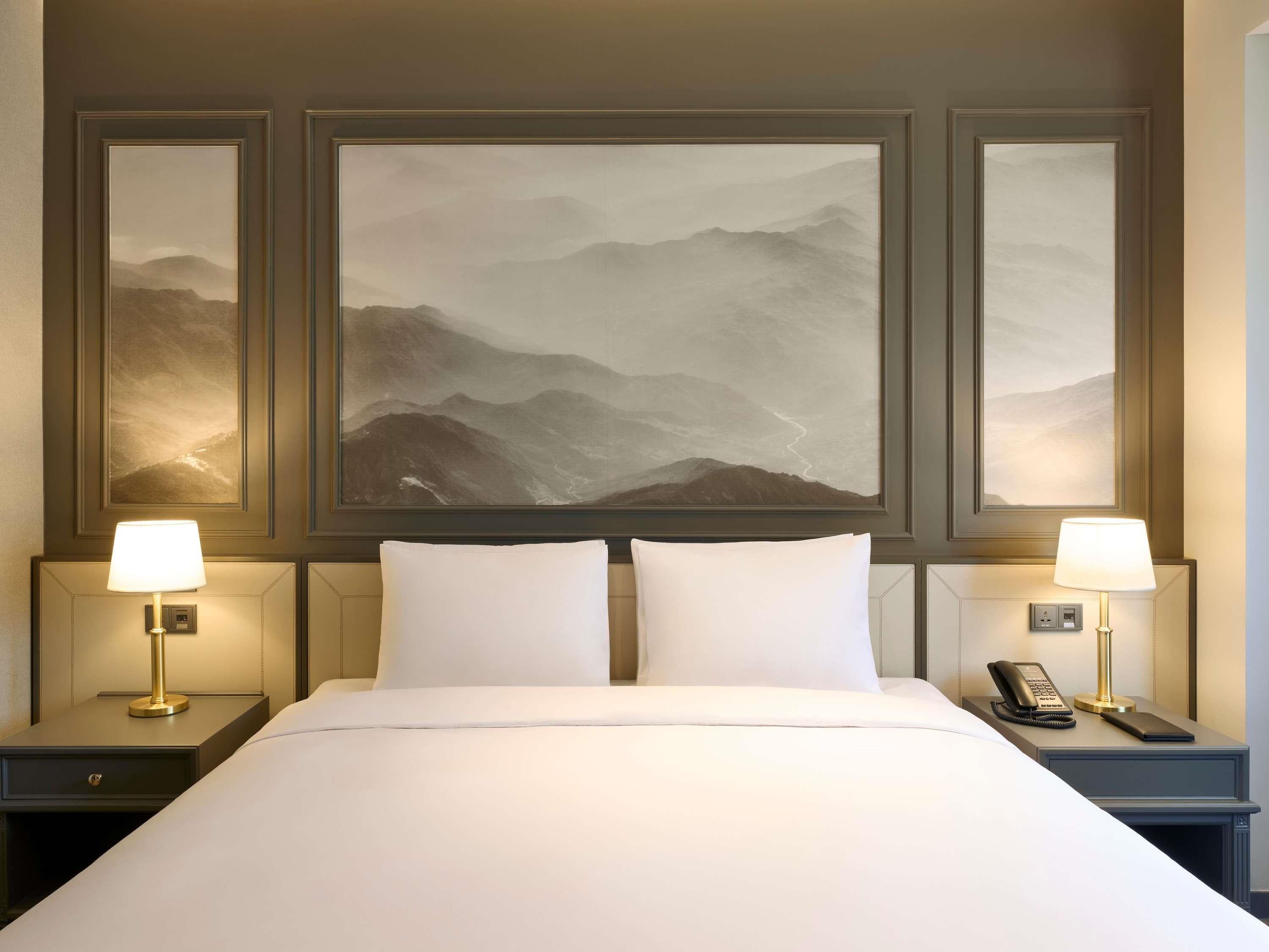 A hotel room with a large bed, two bedside lamps, a telephone, and three framed landscape paintings on the wall.