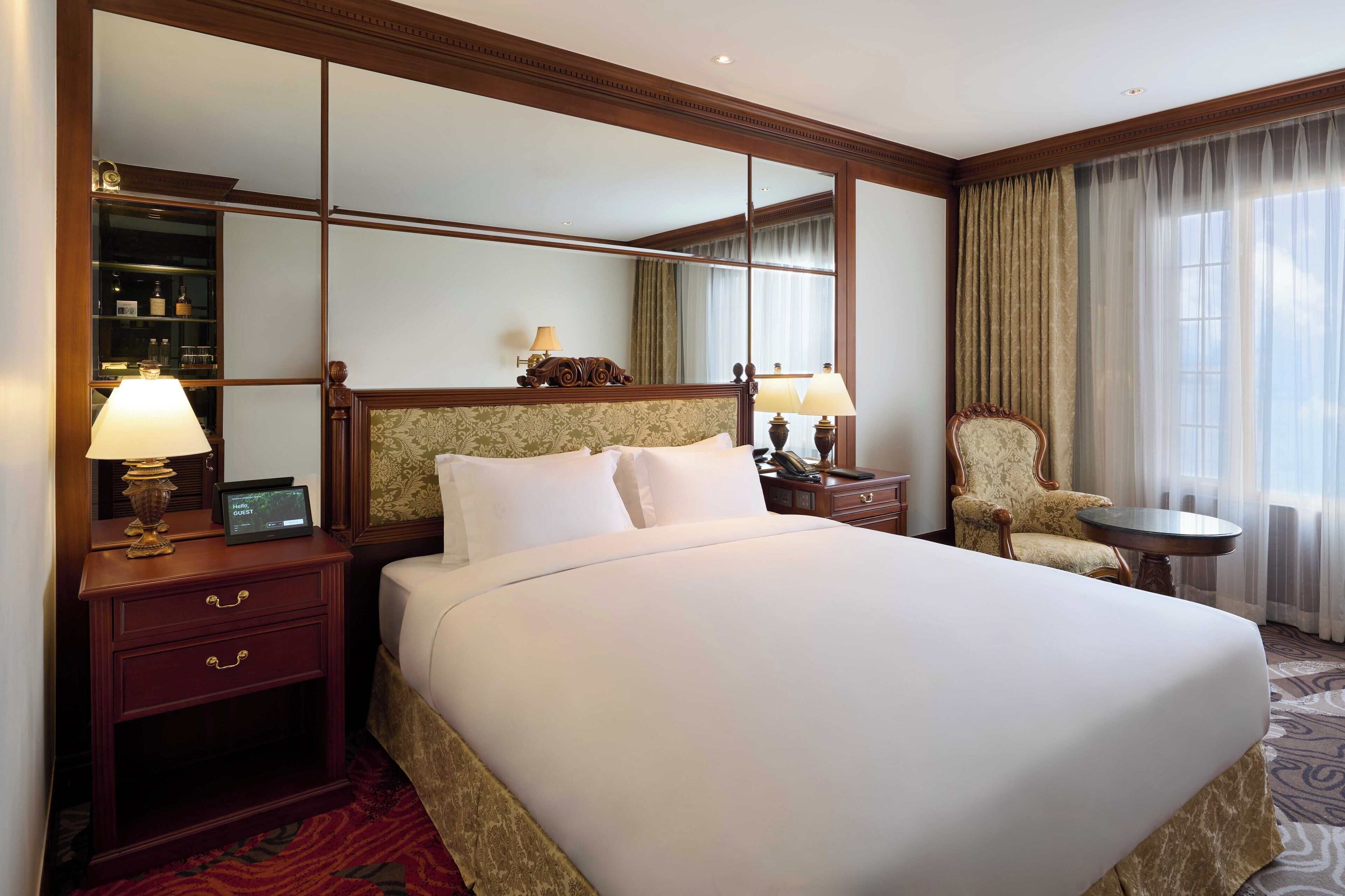 A hotel room with a large bed, a nightstand with a lamp, a chair, a small table, and a mirror.