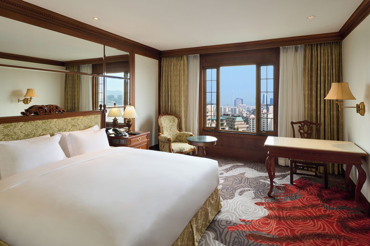A hotel room with a large bed, two chairs, a small table, a desk, a lamp, a mirror, and a view of the city.