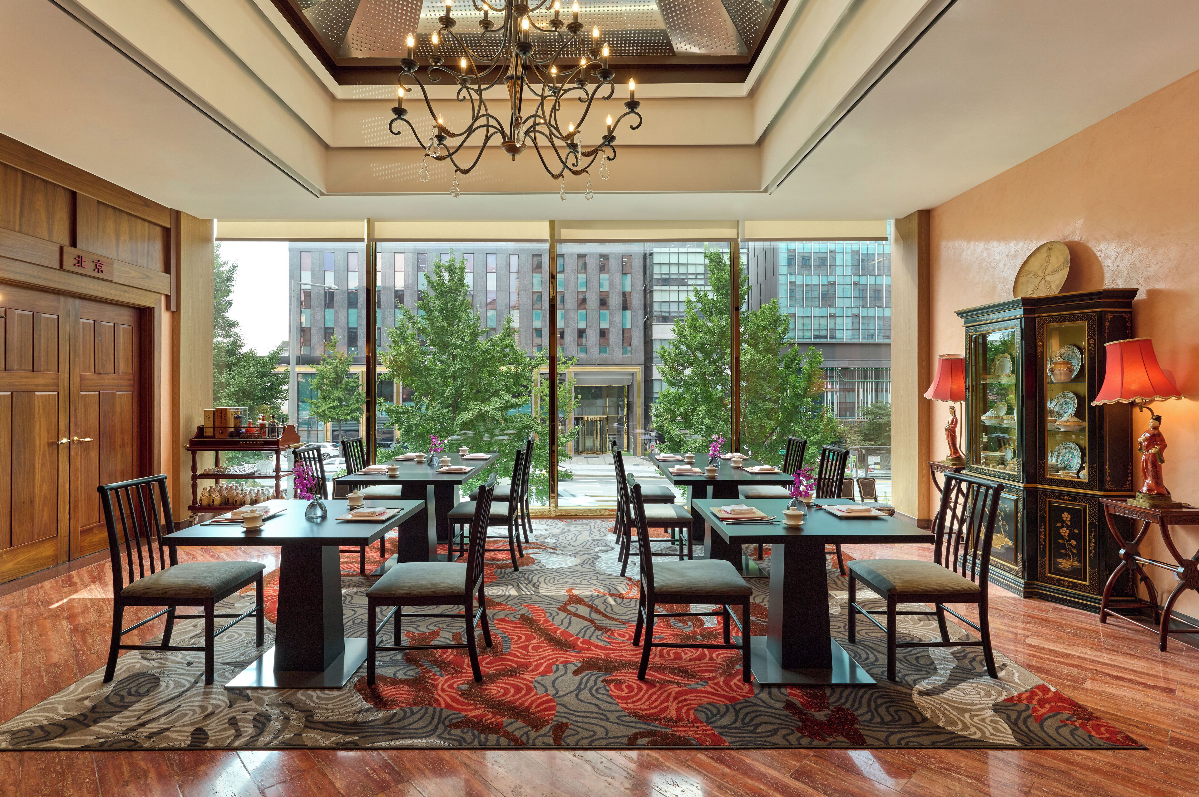 A spacious dining area with multiple tables and chairs, large windows offering a view of the cityscape, and a chandelier hanging from the ceiling.