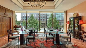 A spacious dining area with multiple tables and chairs, large windows offering a view of the cityscape, and a chandelier hanging from the ceiling.