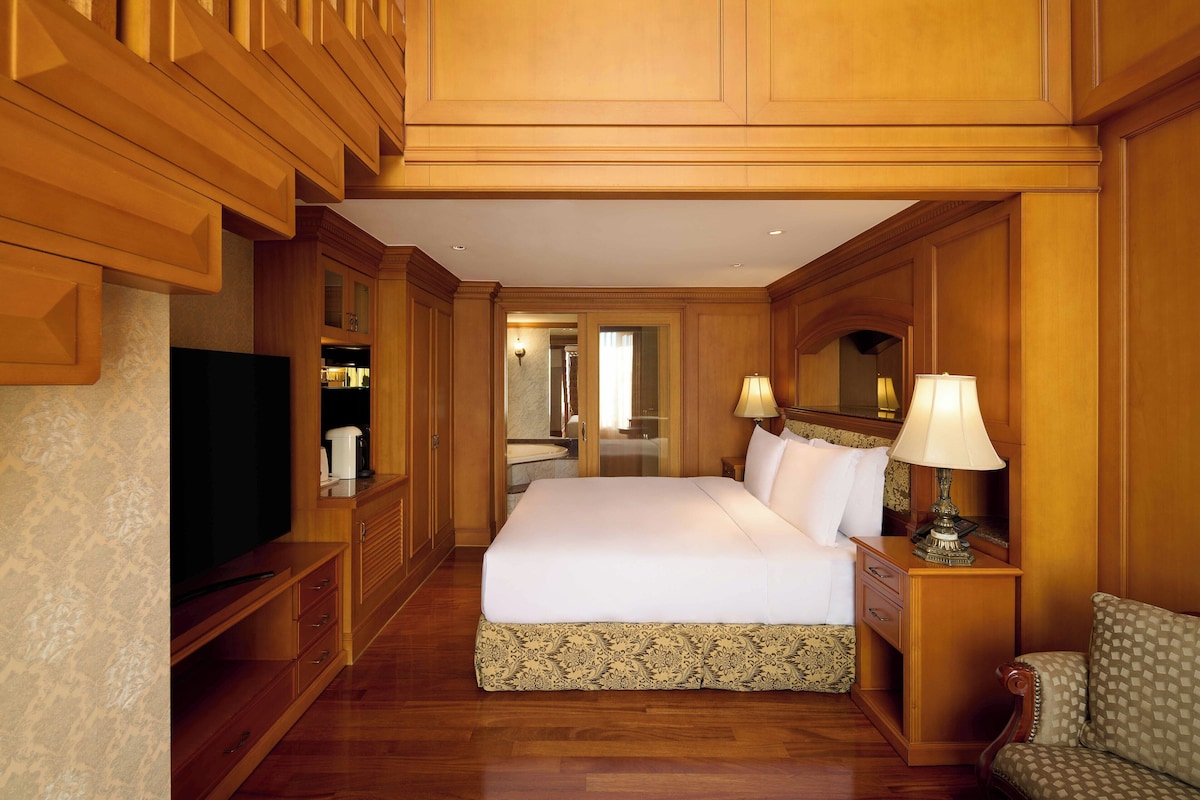 A hotel room with a large bed, wooden paneling, a television, a bedside table with lamps, and a view into the bathroom.