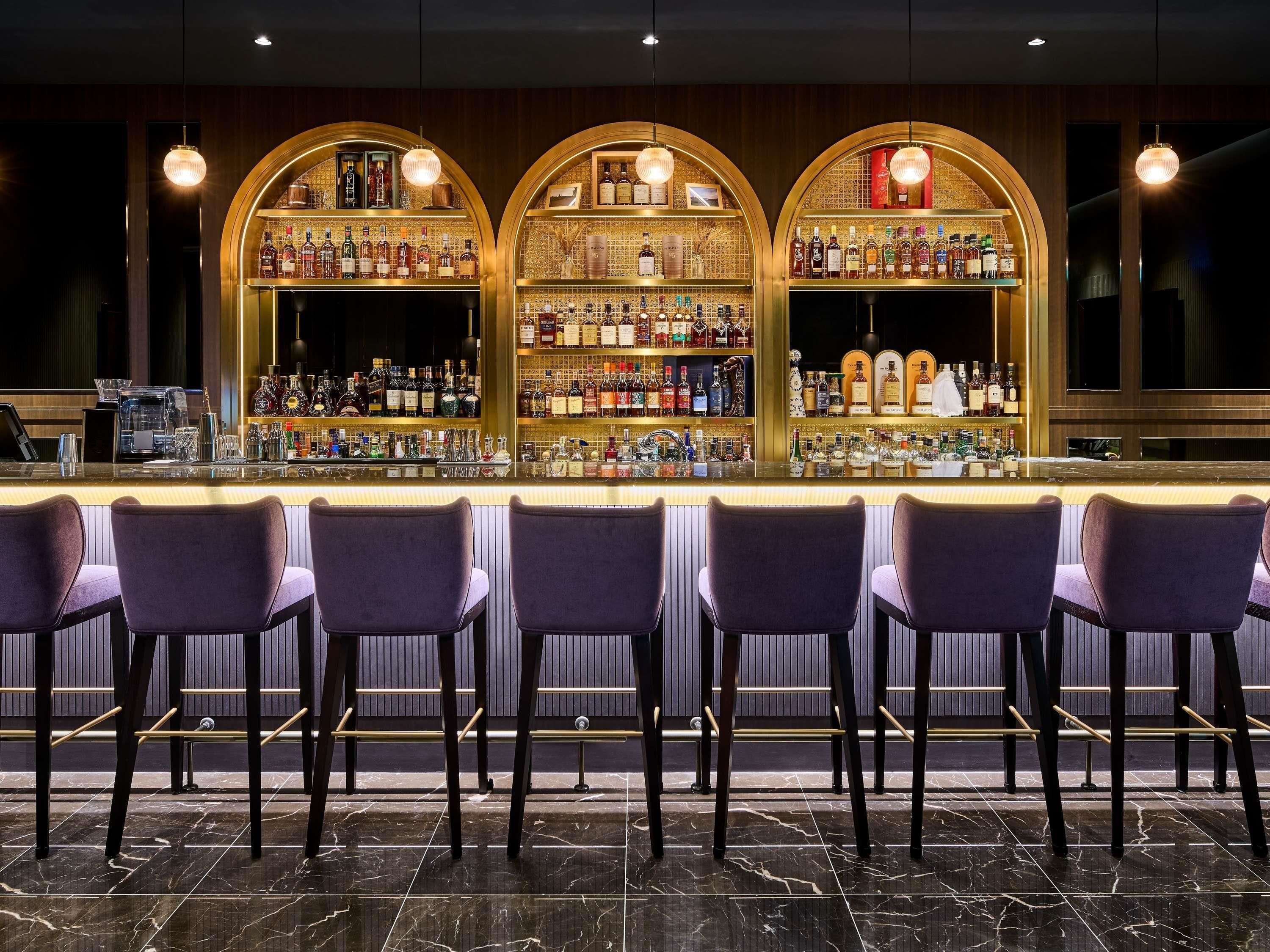 Zona de bar con suelo de mármol, varias estanterías llenas de botellas de licores y taburetes de bar morados.