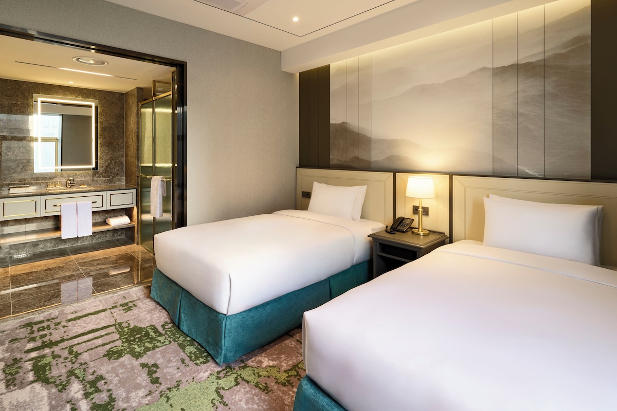 A hotel room with two beds, a bathroom with a large mirror, and a wall-mounted artwork.