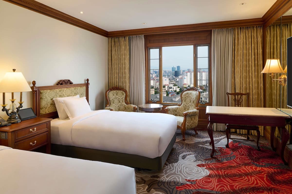A hotel room with a large bed, a desk, a chair, a small table, and a view of the city through large windows.