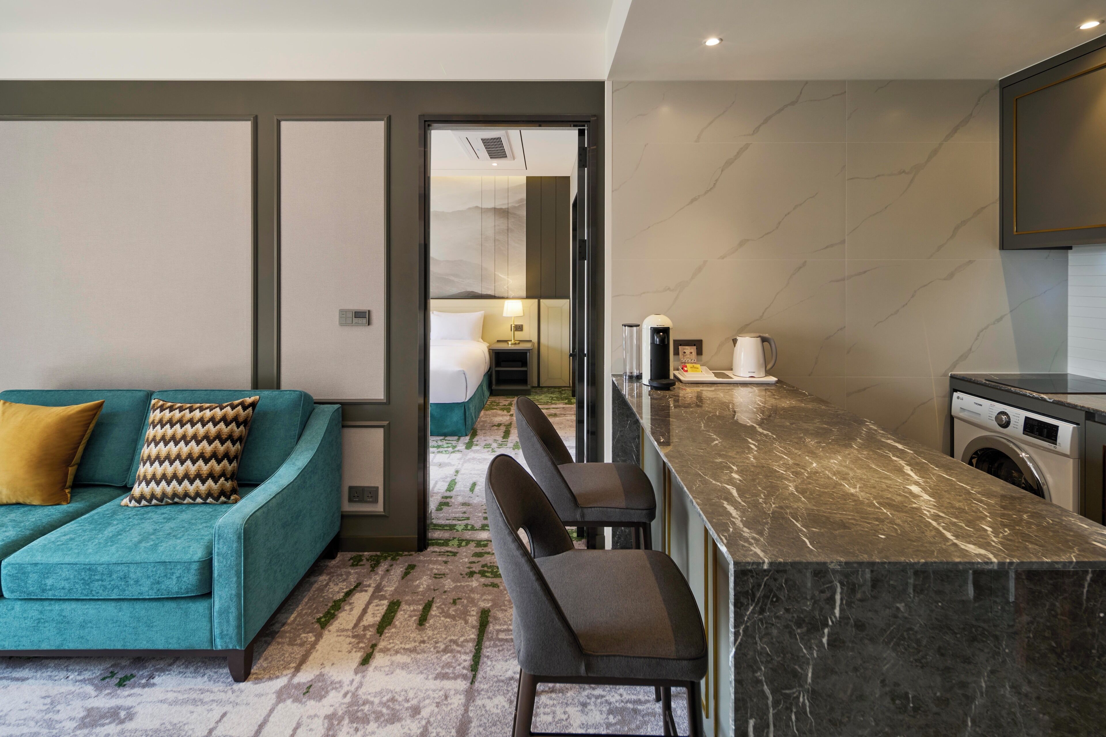 Quarto de hotel moderno com sofá verde-água, ilha de cozinha em mármore e máquina de lavar roupa.