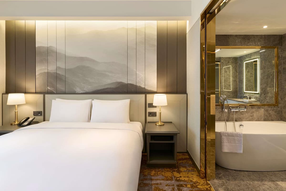 A hotel room with a bed, two bedside lamps, a nightstand, a bathroom with a bathtub, and a large mirror.
