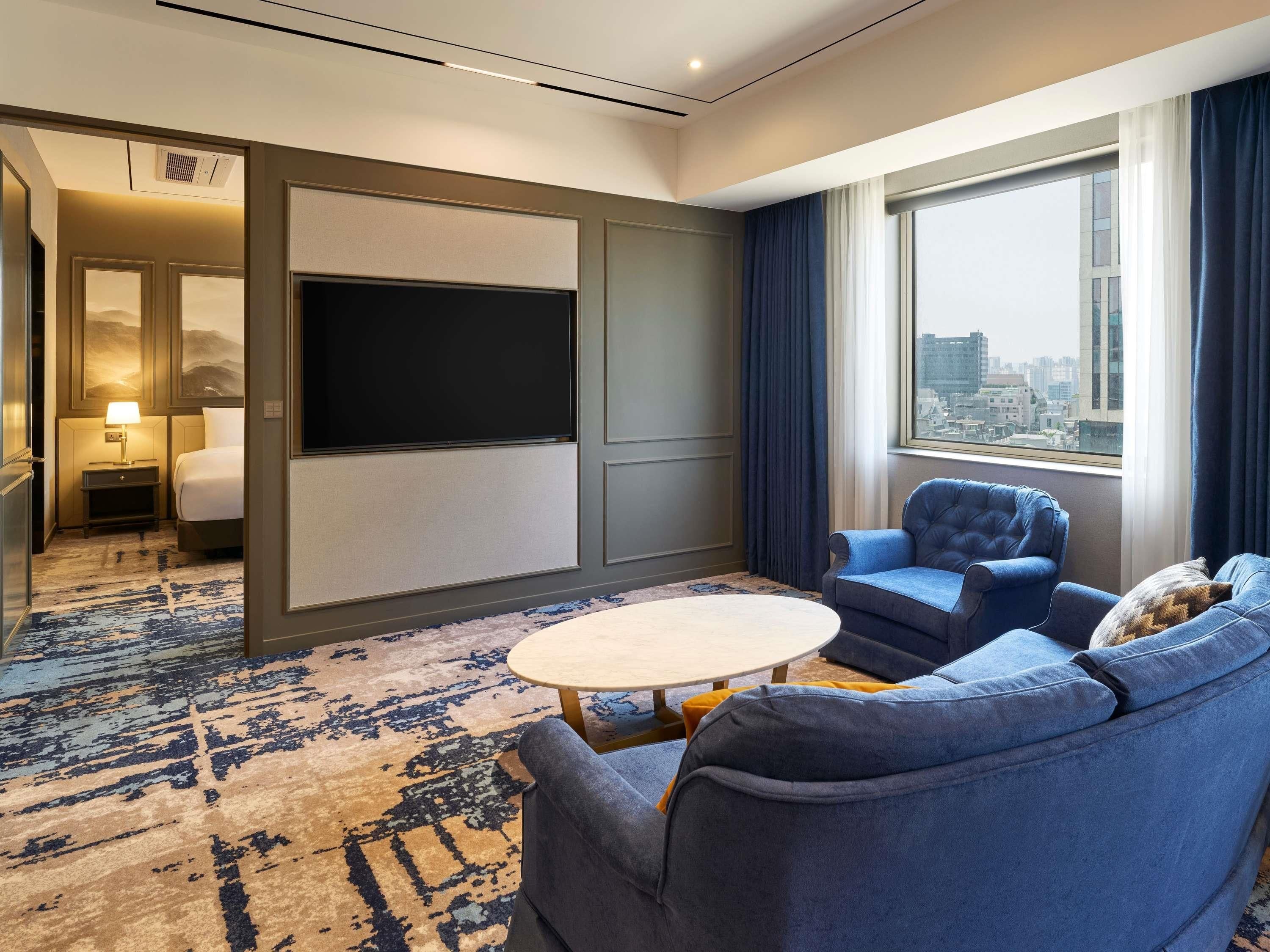 Habitación de hotel con una cama grande, un televisor de pantalla plana, una zona de estar con sofá y sillones, una mesita pequeña y vistas a la ciudad a través de un ventanal amplio.
