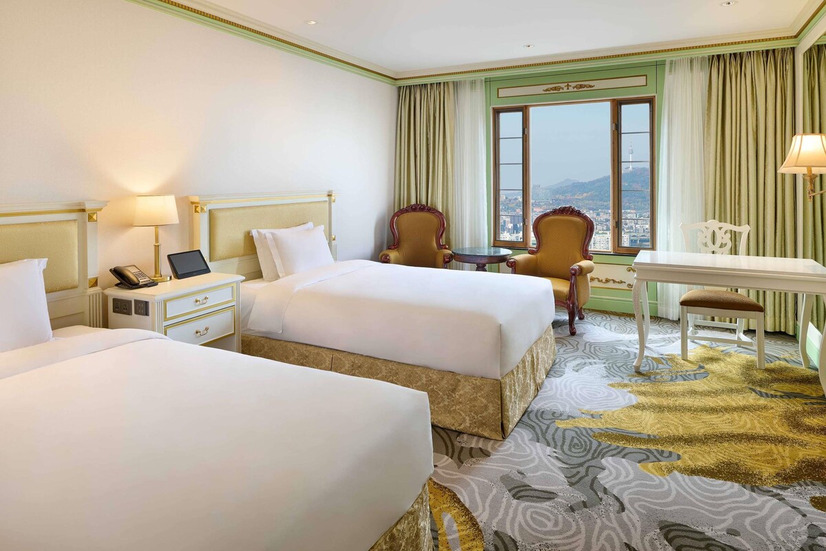 A hotel room with two beds, a desk, a chair, a phone, and a view of the city.