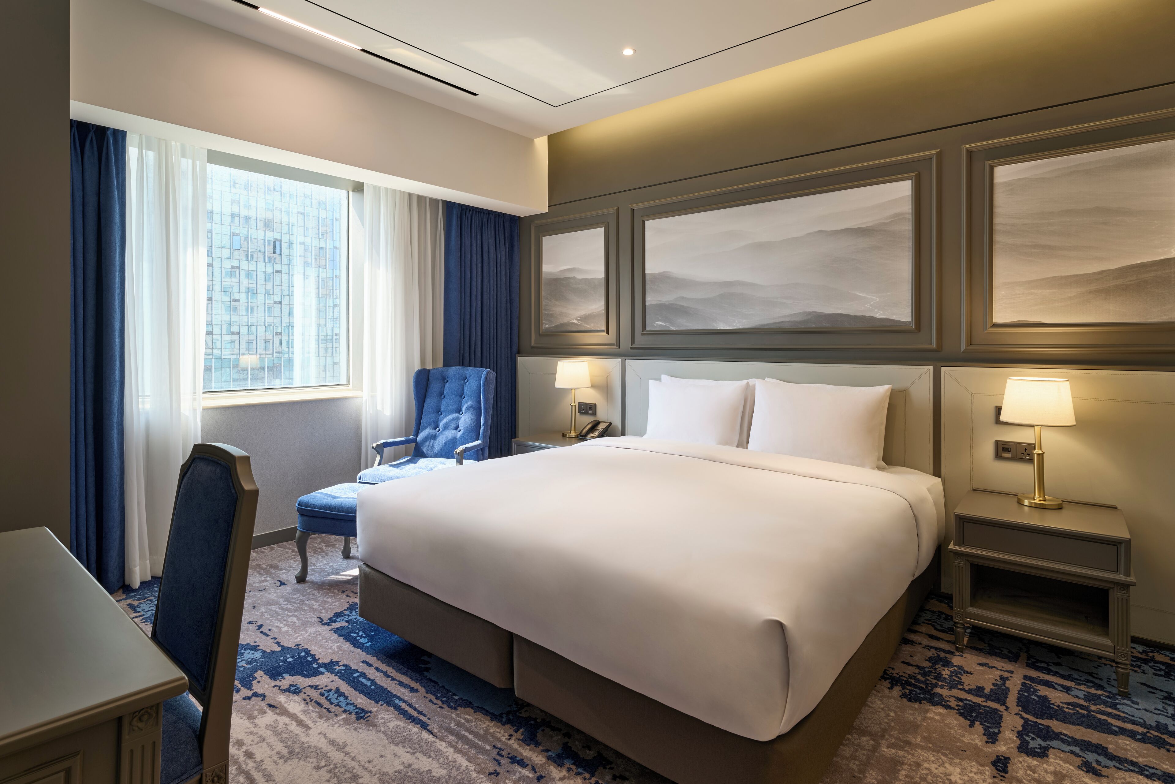 A hotel room with a large bed, a desk, a chair, a window with curtains, and artwork on the walls.