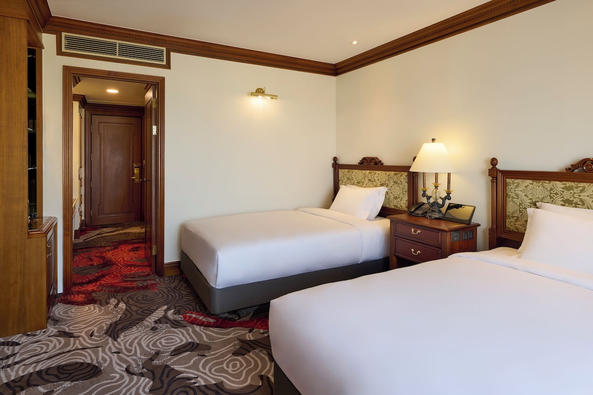A hotel room with two beds, a wooden nightstand with a lamp, a telephone, and a red carpeted hallway.