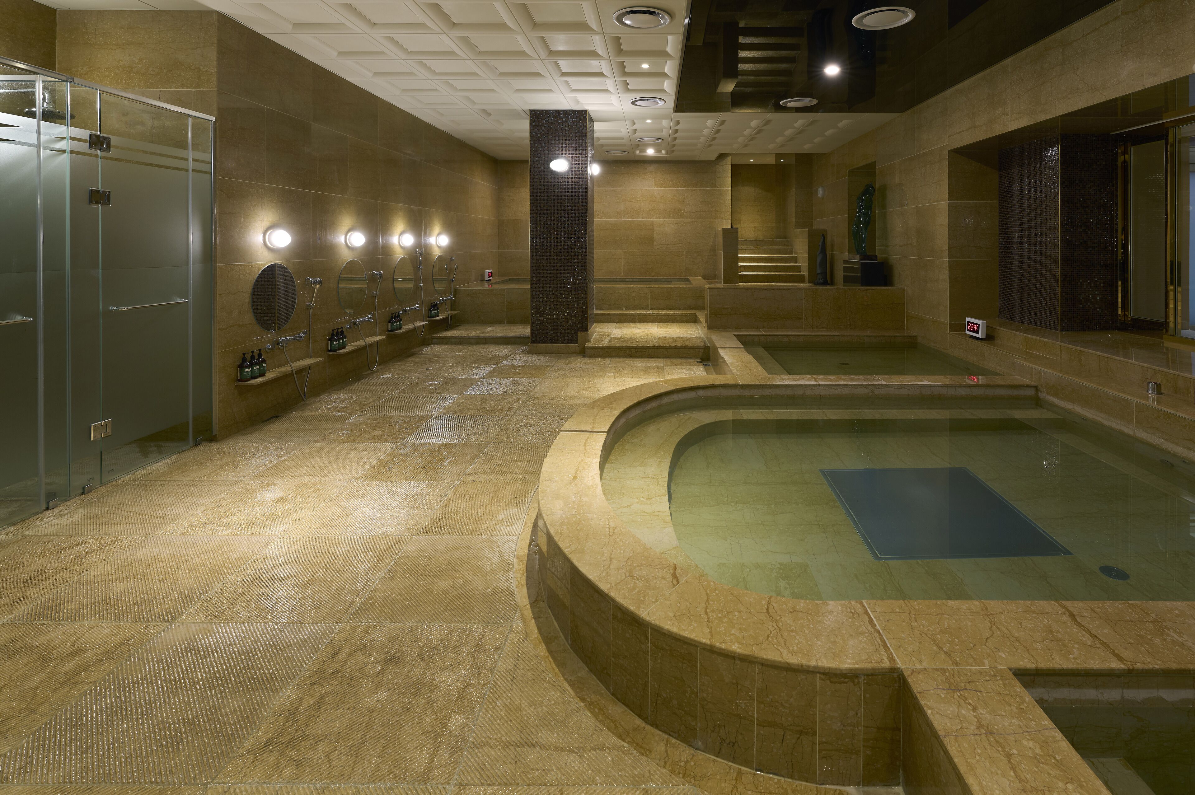 Un espace piscine intérieure moderne, doté de plusieurs douches, d’une piscine centrale et d’un sol carrelé en pierre.