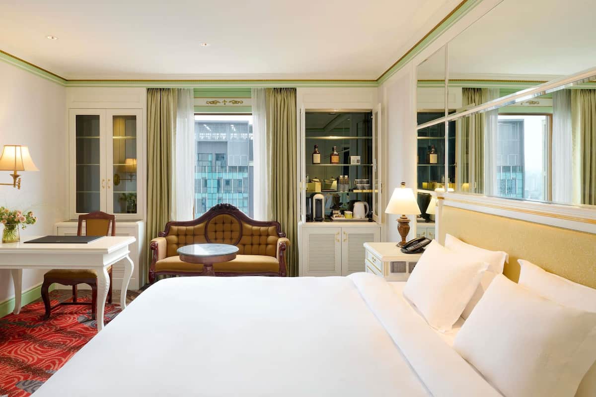 A hotel room with a large bed, a desk with a chair, a small round table, a sofa, and a large mirror.