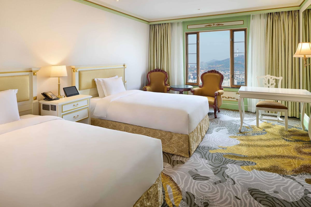 A hotel room with two beds, a desk, a chair, a phone, and a view of the city.
