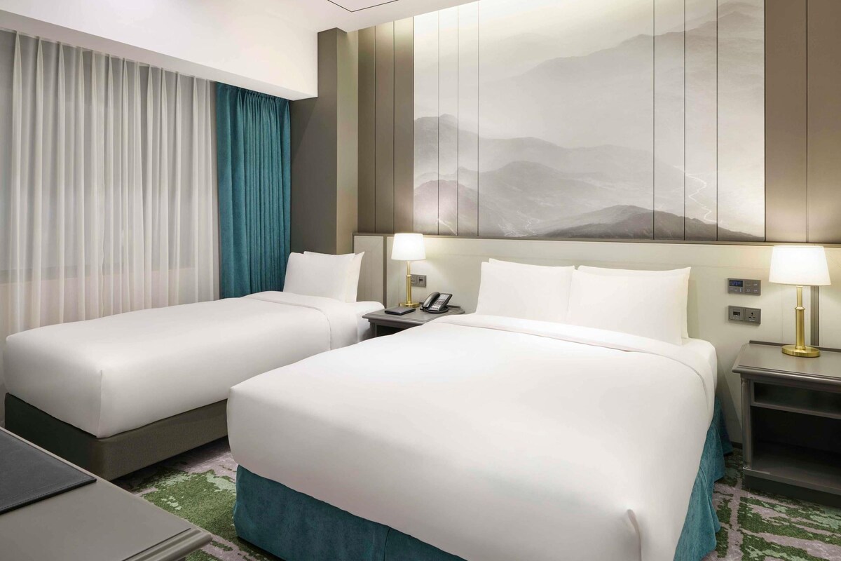 A hotel room with two beds, a desk, a phone, and a painting on the wall.