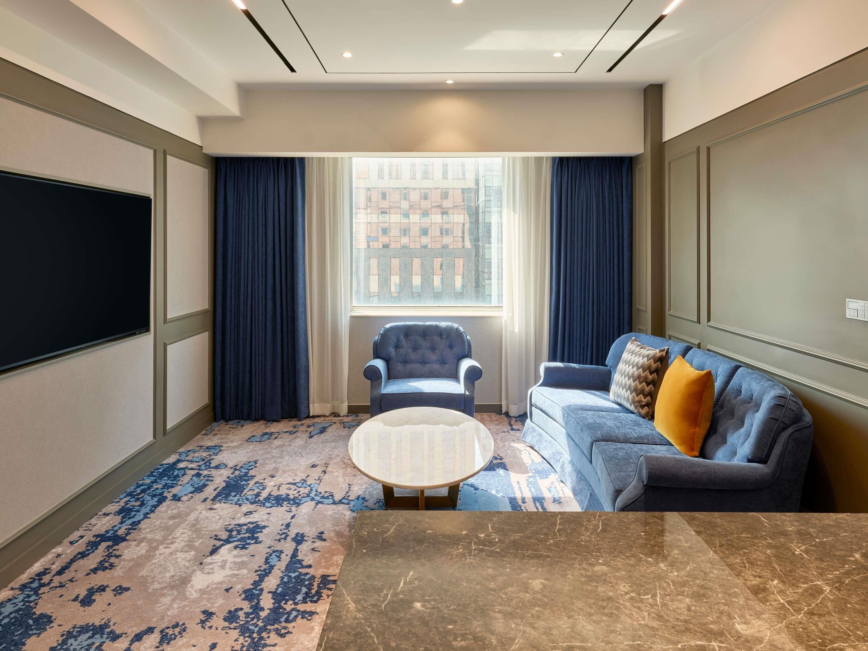 A hotel room with a large window, a blue armchair, a blue sofa, a marble coffee table, and a flat-screen TV.