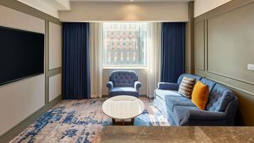 A hotel room with a large window, a blue armchair, a blue sofa, a marble coffee table, and a flat-screen TV.