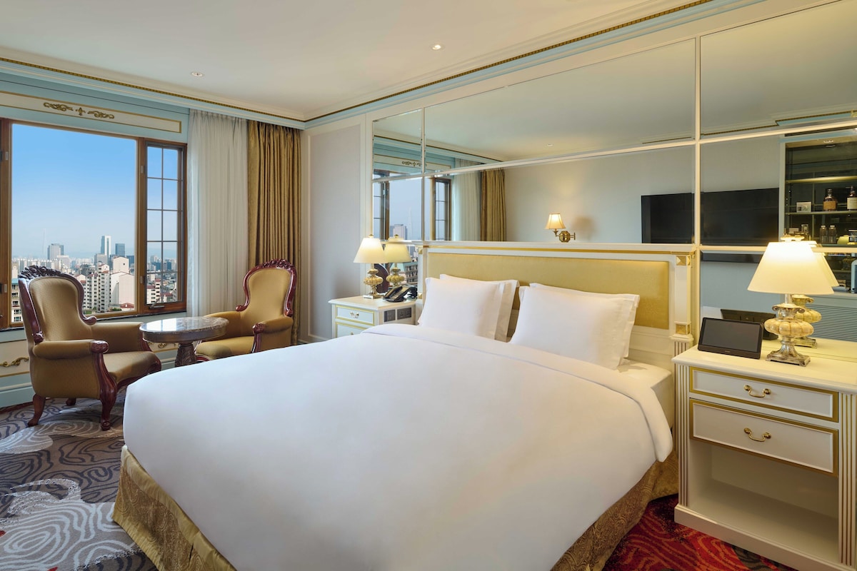 A hotel room with a large bed, a round table, two armchairs, a nightstand with a lamp, a TV, and a city view through the window.