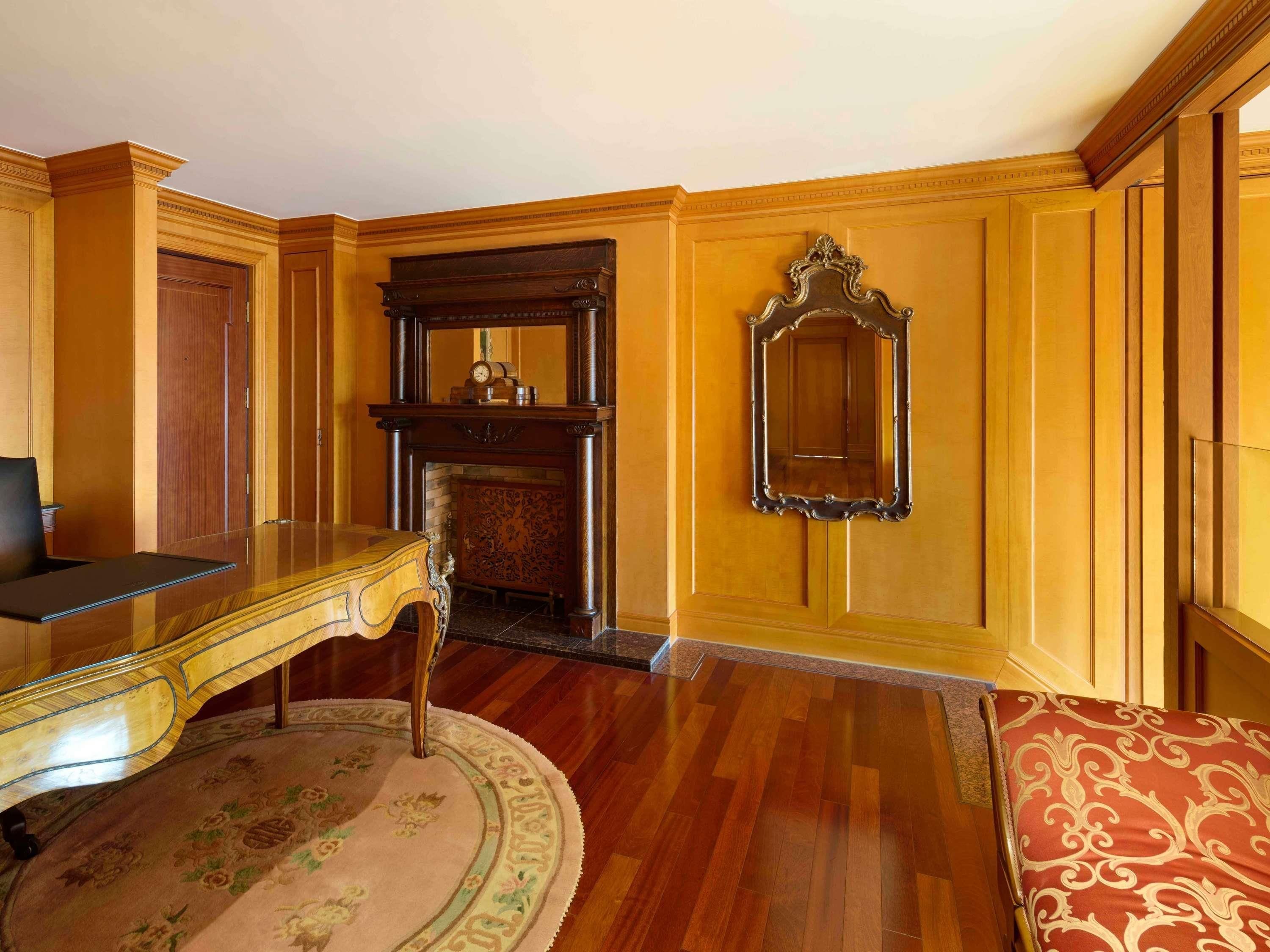 A room with a wooden floor, a glass-top desk, a patterned rug, a fireplace, and a mirror on the wall.