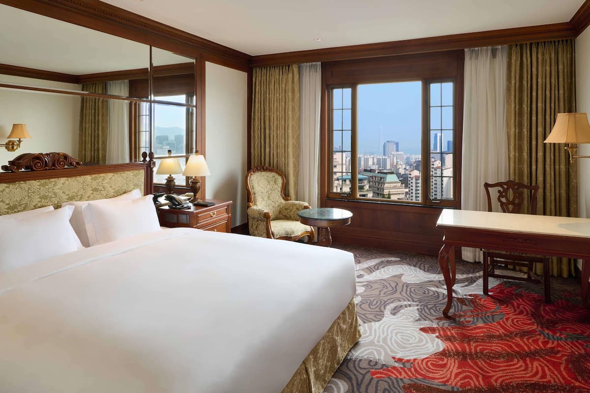A hotel room with a large bed, a desk, a chair, a small table, a mirror, and a city view through large windows.