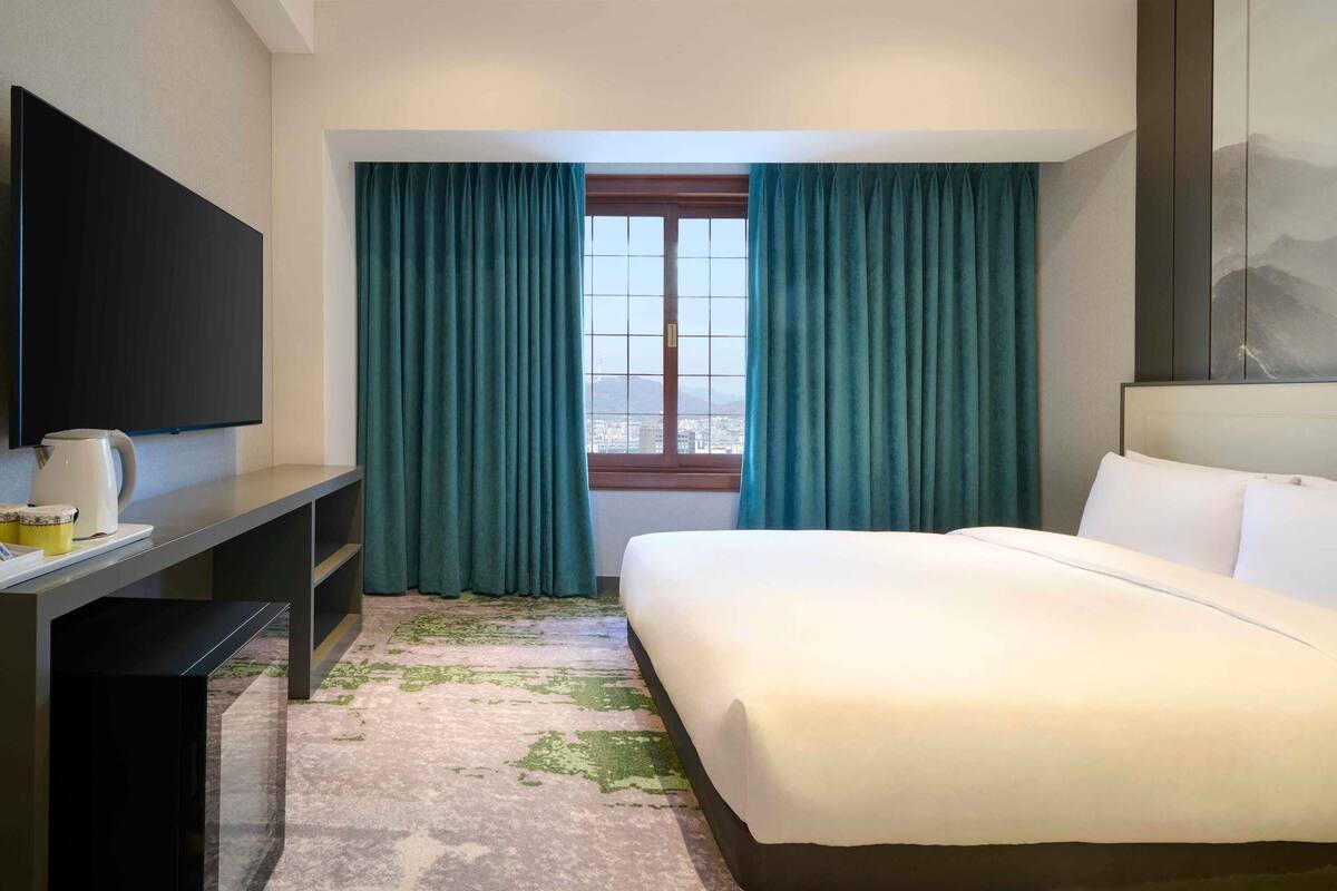 A hotel room with a bed, a desk with a flat-screen TV, a window with curtains, and a view of the cityscape.