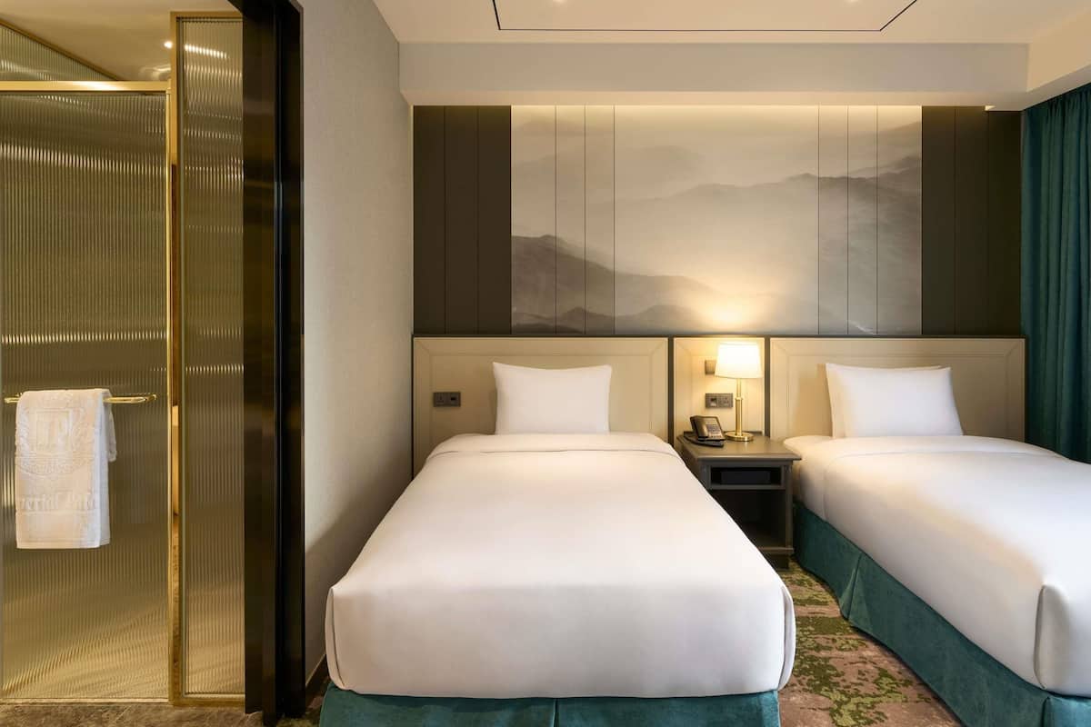 A hotel room with two beds, a shower area, a nightstand with a lamp, and a wall-mounted artwork.