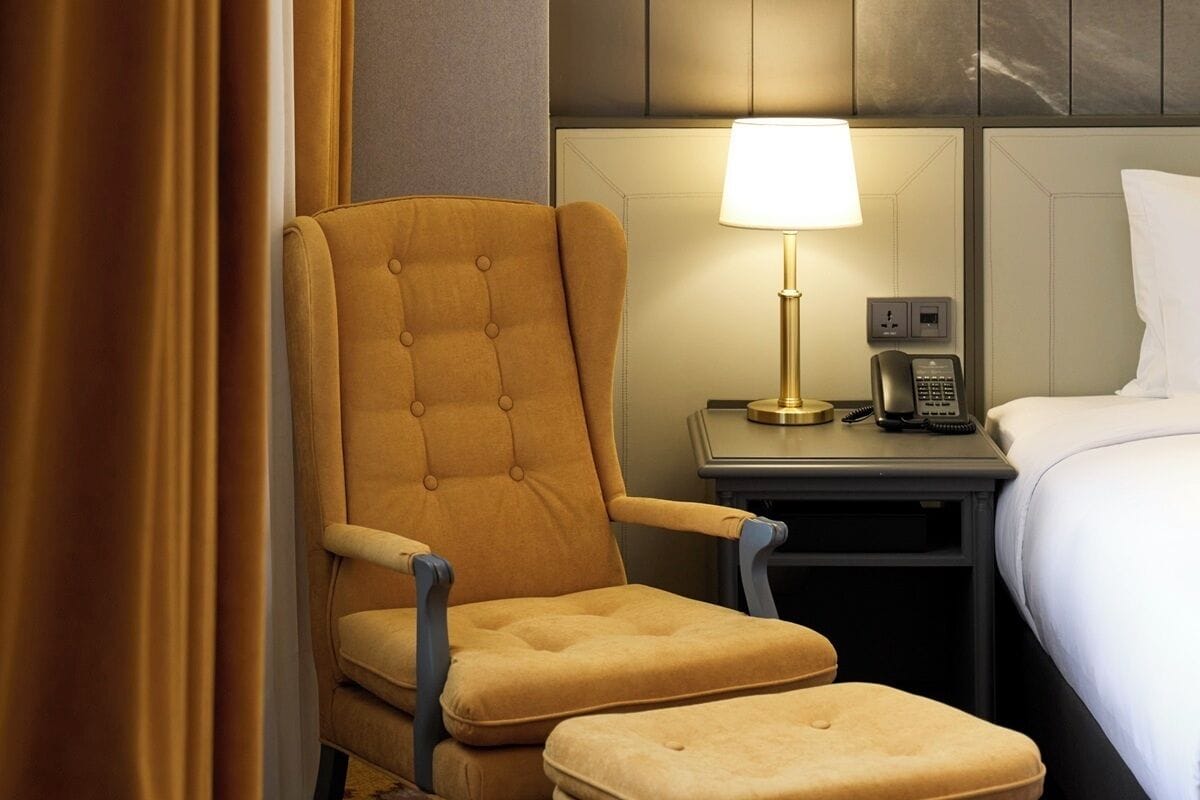 A hotel room with a tufted armchair, a bedside table with a lamp, a telephone, and a bed with white bedding.