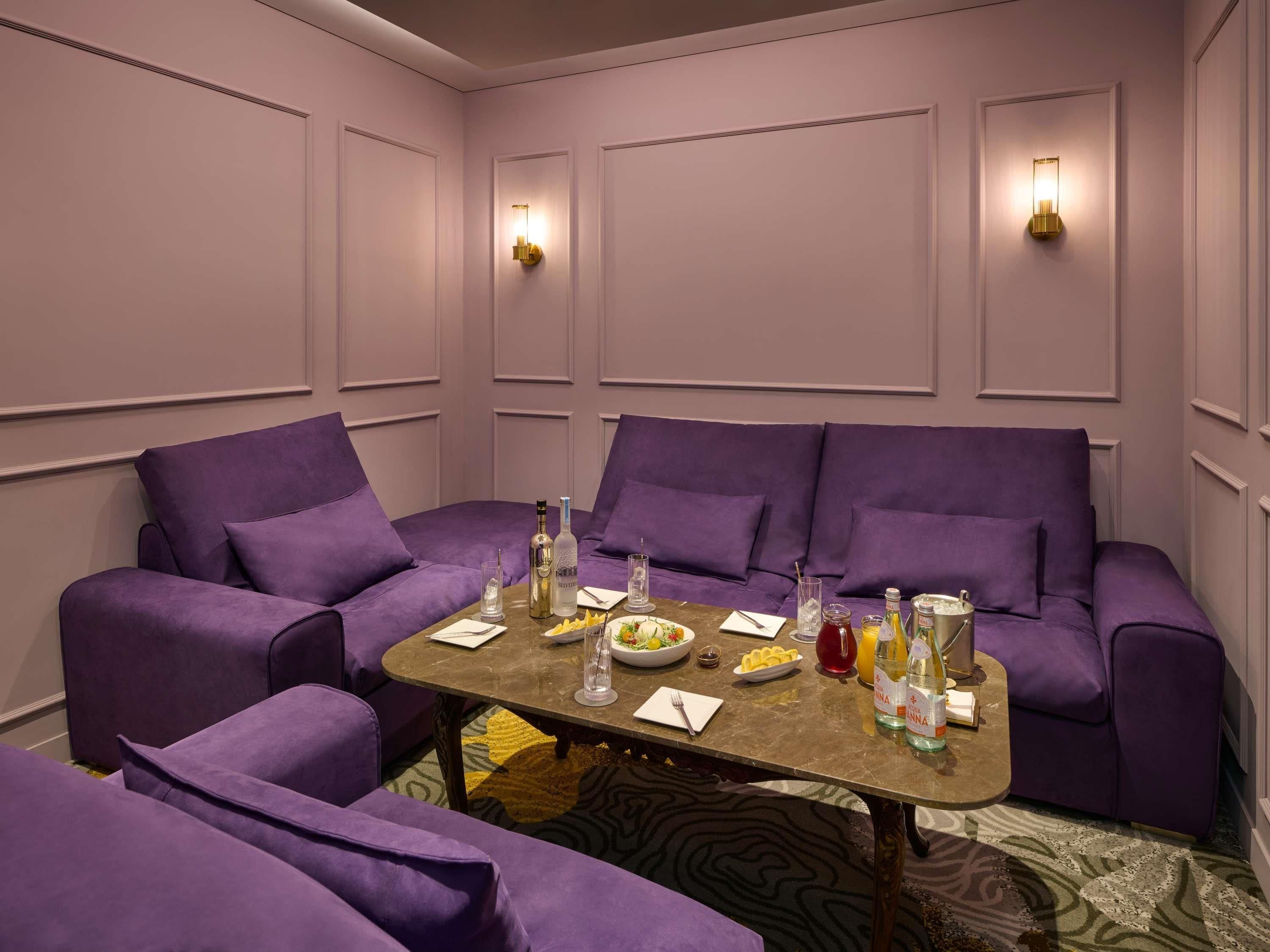 Una habitación con un sofá morado, una mesa de madera con ensalada, bebidas y una botella de agua.