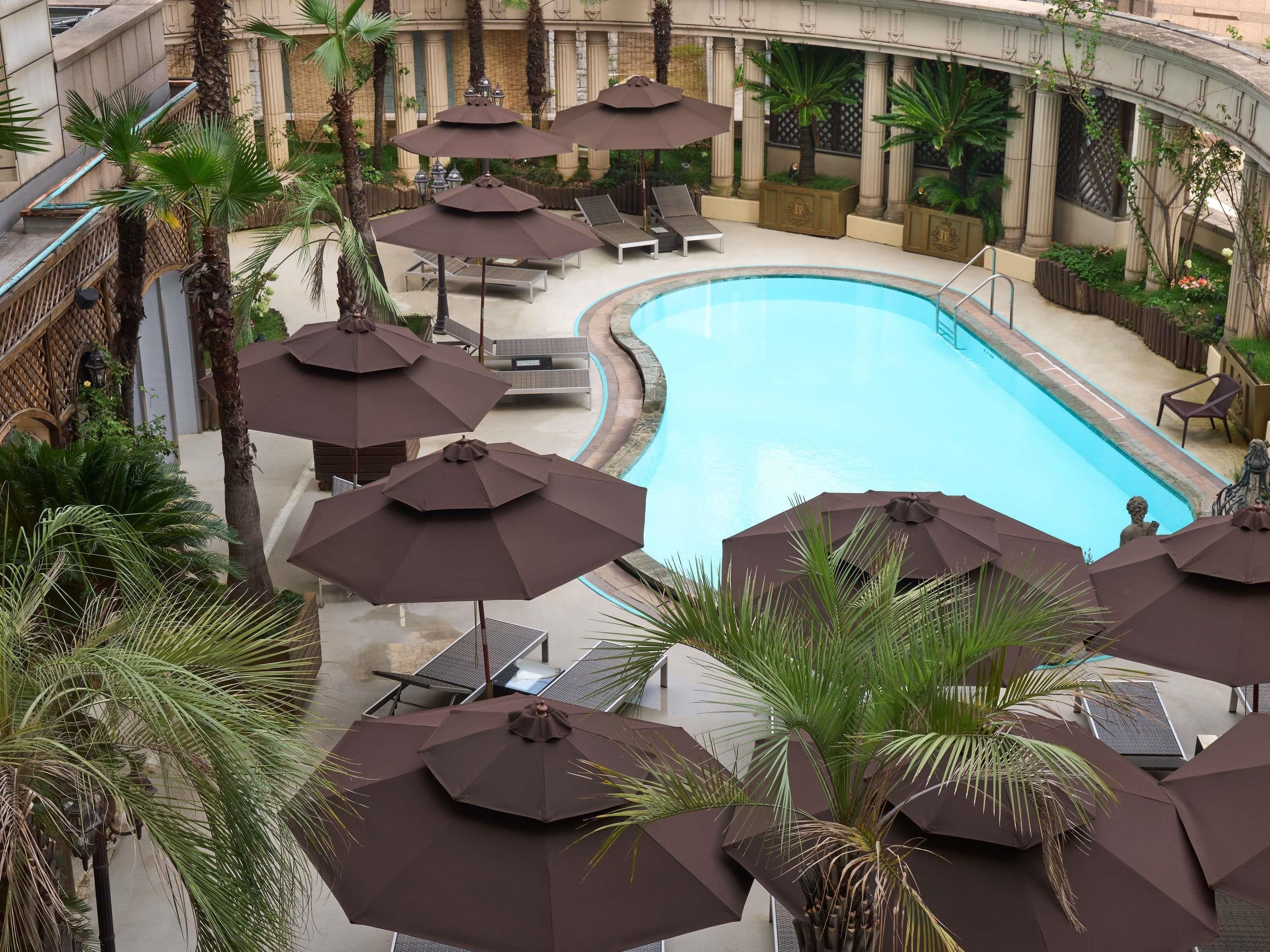 A swimming pool with multiple umbrellas and lounge chairs, surrounded by palm trees and architectural columns.