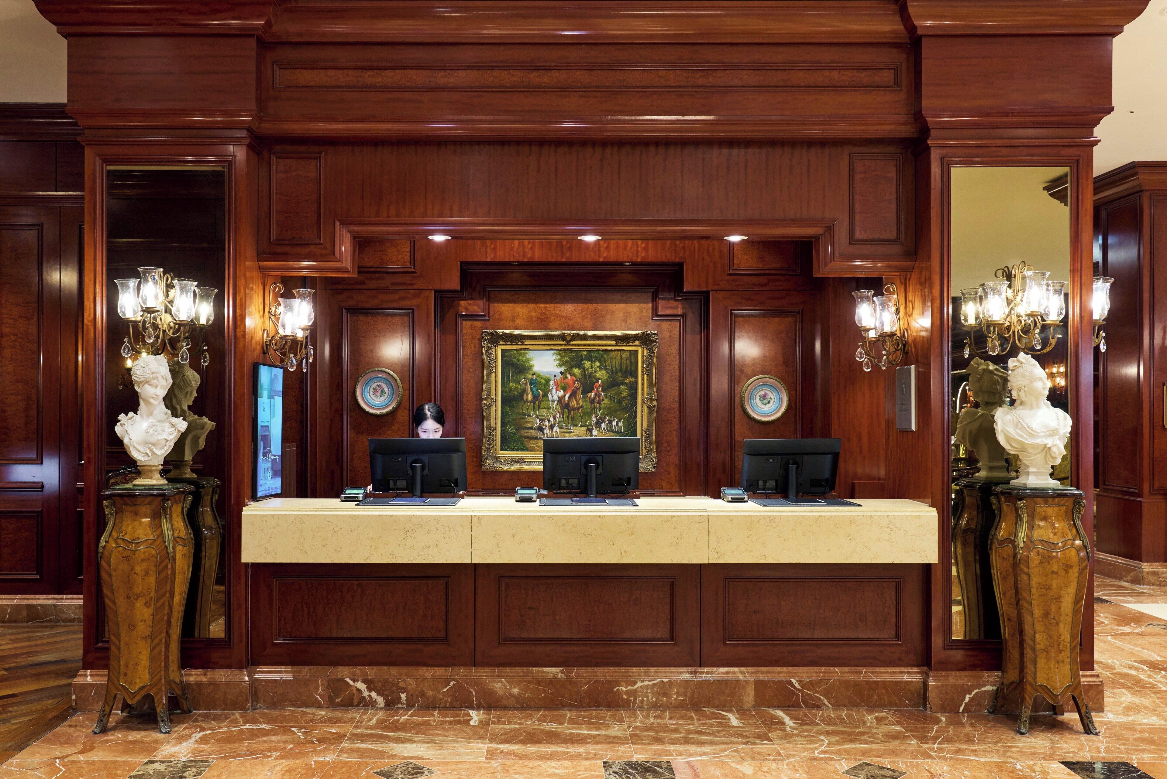 A reception desk with two computer monitors, a framed painting above, and two ornate vases on either side.