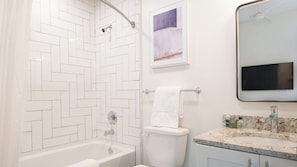 A bathroom with a white herringbone tile shower, a toilet with a towel, a framed picture, and a sink with a marble countertop.