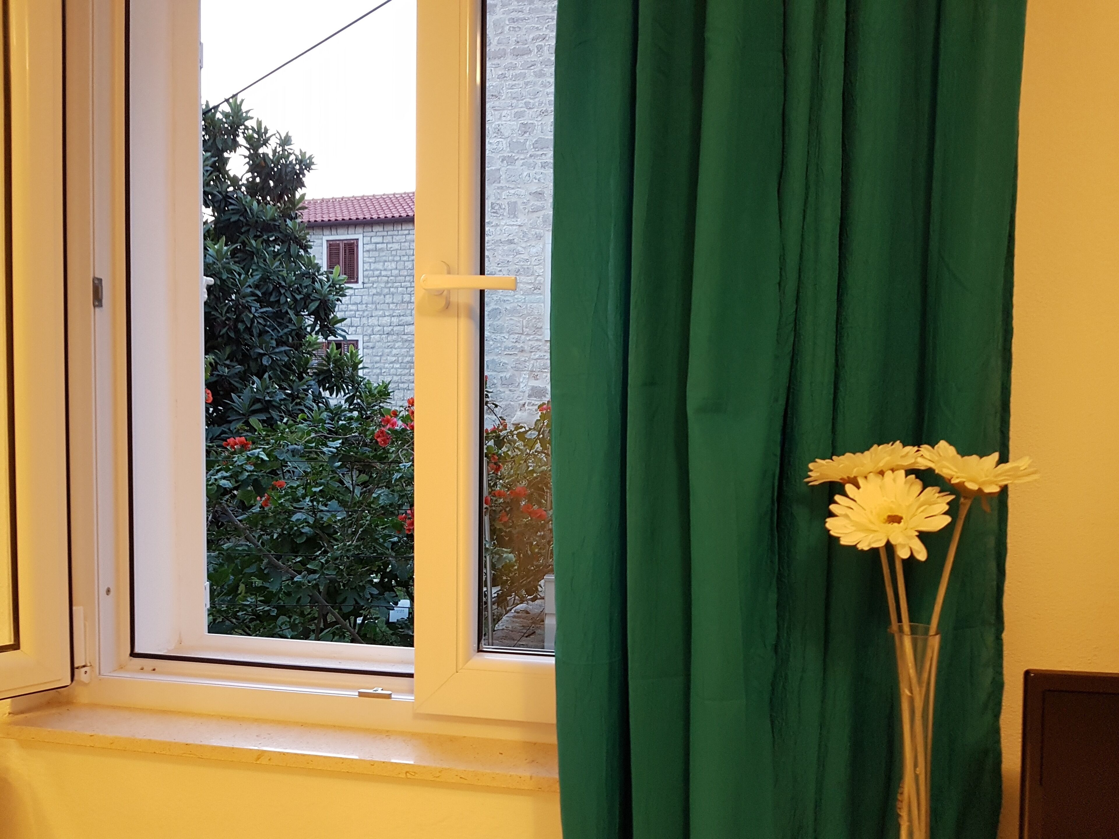 A window with a green curtain, a white vase with flowers, and a view of a building with a red roof.
