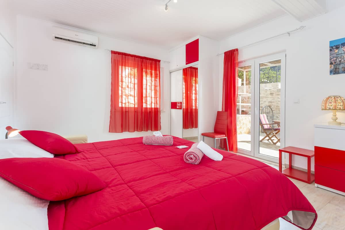 Une chambre avec une couvre-lit rouge, un climatiseur blanc, une chaise rouge et une vue sur l’extérieur par une porte vitrée.